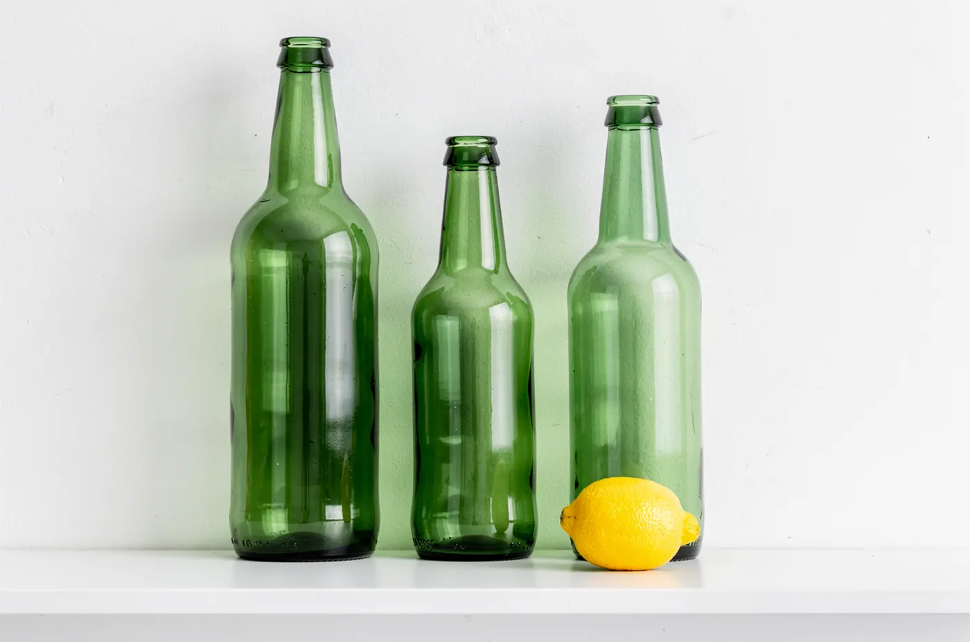 FLUX.2 Pro: Exactly three green glass bottles standing on a white shelf. The tallest bottle on the left, the shortest in the middle, and a single yellow lemon on the shelf in front of the right-hand bottle. Plain white wall, soft even light.