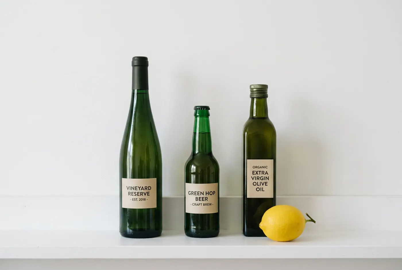 Nano Banana 2: Exactly three green glass bottles standing on a white shelf. The tallest bottle on the left, the shortest in the middle, and a single yellow lemon on the shelf in front of the right-hand bottle. Plain white wall, soft even light.