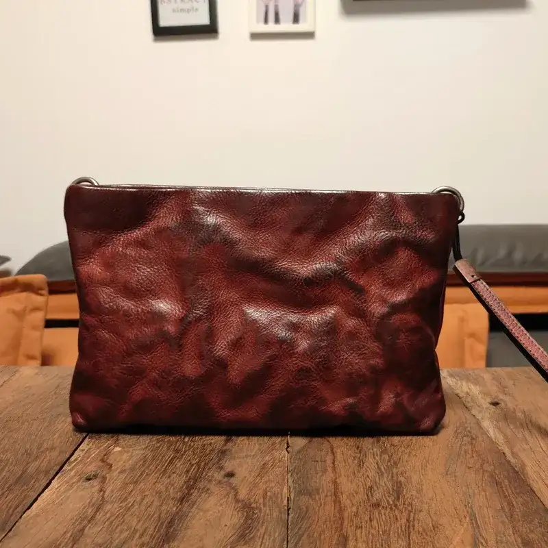 A maroon leather clutch standing on a reclaimed-wood table in a living room, under a warm lamp with no fill light, an orange armchair and a grey sofa behind it