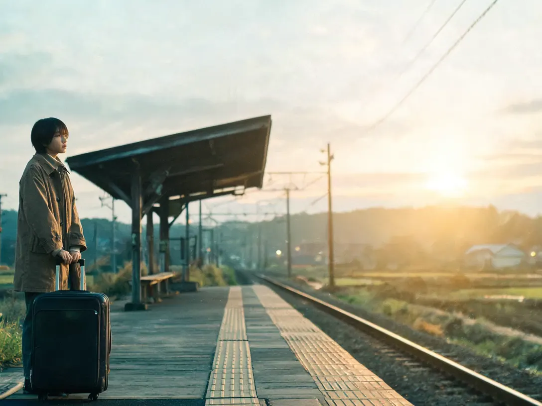 A traveller with a suitcase on a rural Japanese platform at dusk as a train pulls in and stops, taking one deep breath before she boards